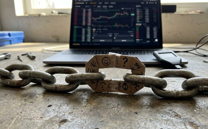 A cardboard link marked with a smiley face and question marks connecting two rusty metal chains on a workbench, with a laptop displaying trading charts in the background.