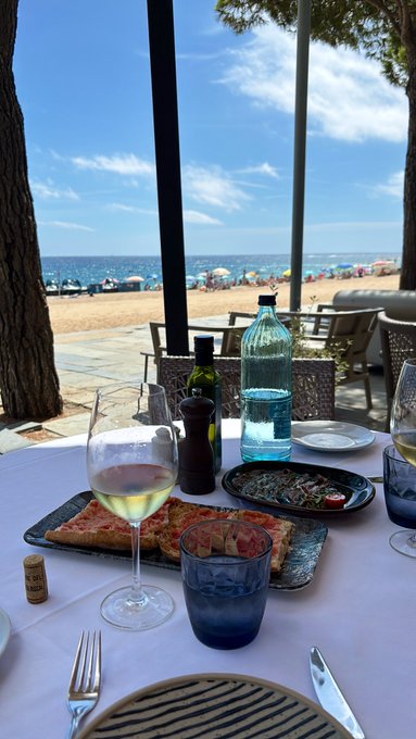 A table set with pizza, wine, and water overlooks a sunny beach scene with blue water and umbrellas.
