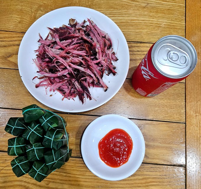 A wooden table featuring a white plate with shredded purple/pink meat, a can of Coca-Cola, a small plate with red sauce, and a cluster of leaf-wrapped parcels tied with string.