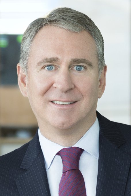 A professional headshot of a smiling man with gray hair and blue eyes, wearing a dark pinstripe suit, white shirt, and red patterned tie.
