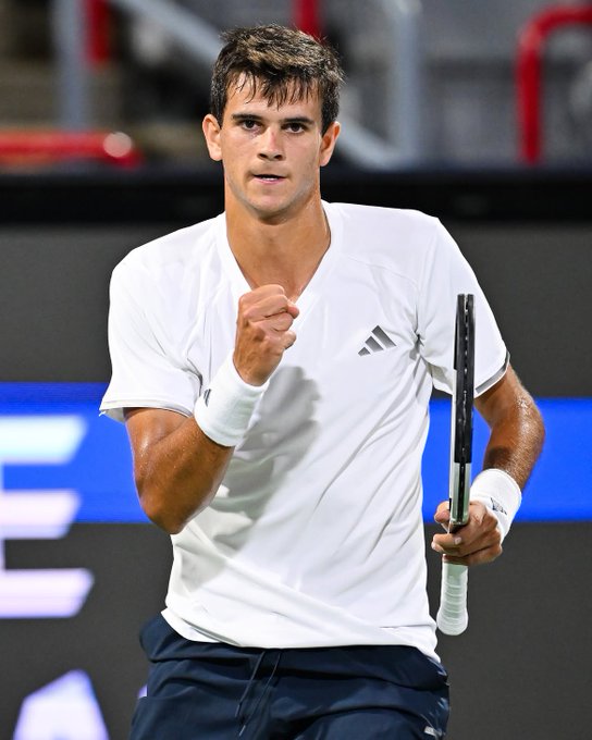 A male tennis player in a white Adidas shirt and navy shorts celebrates with a clenched fist while holding a tennis racket on a court.
