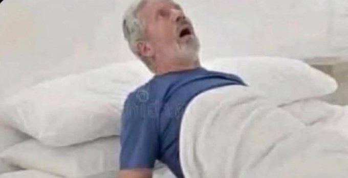 A man with gray hair sleeps soundly in a white bed, his head resting on pillows with his mouth open.