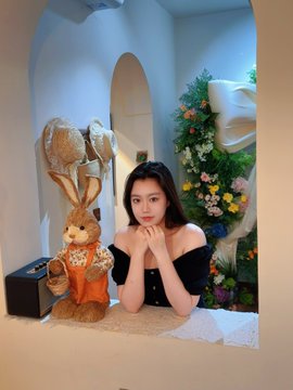 The image features a woman sitting at a desk with a stuffed rabbit next to her, both of them positioned near an archway or doorway. The background includes a wall clock and some flowers, adding to the overall atmosphere of the scene. The photo appears to be related to a post on social media, as indicated by the caption "昨天对着OPENAI/USDT纠结了半天，想入场又怕踩进来就踏空 蒜鸟蒜鸟".