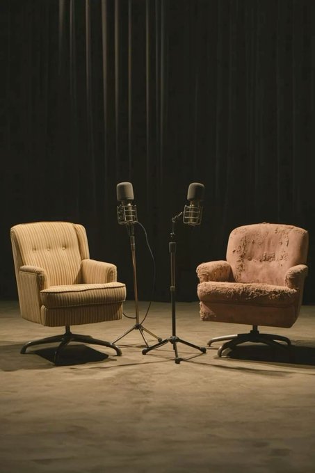 A vintage-style photograph showing two worn, mid-century armchairs—one striped and one pink and shabby—facing each other with two microphones on stands set up between them on a stage with a dark curtain backdrop.