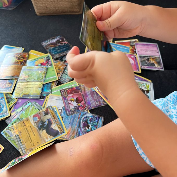 A child's hands sorting through a large, colorful pile of Pokemon trading cards spread out on a dark surface.