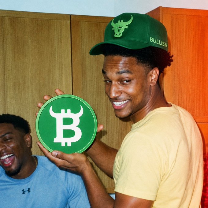 A smiling young man wearing a green cap and shirt holds a green circular sign with the Bitcoin 'B' logo, with another person laughing beside him.