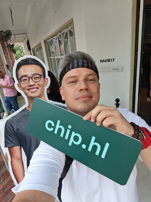 A man wearing a headband holds a green 'chip.hl' sign while posing with a cardboard cutout of a smiling young man with glasses.