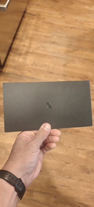 A person's hand holding a small dark gray card or envelope with an 'X' symbol embossed in the center, with a wooden floor in the background.