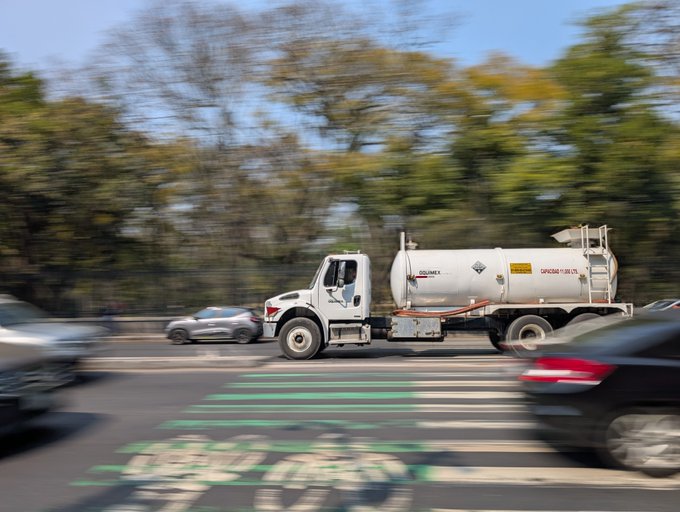 A white tanker truck with hazard placards drives along a tree-lined road, captured with motion blur as a gray SUV and dark sedan travel alongside it.