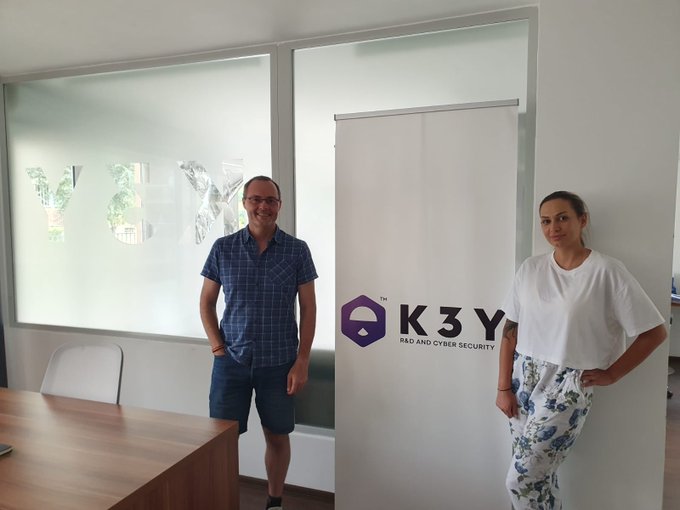 Two people standing in an office next to a banner for 'K3Y - R&D and Cyber Security', with frosted glass windows in the background displaying 'K3Y'.