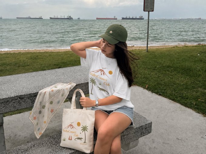 A young woman wearing a green cap and a 'Valley Connect Club' white t-shirt sits on a stone bench by the sea, with cargo ships visible in the background and a matching club tote bag beside her.
