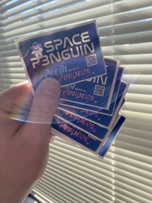 A hand holds a fanned-out stack of holographic stickers that read 'SPACE PENGUIN' and 'Profit With A Purpose', featuring a QR code and a cartoon penguin logo.