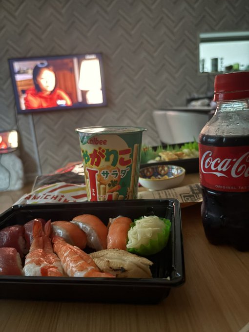 A Japanese meal setup featuring a bento box with various sushi, a cup of Calbee Jagariko salad snack, a bottle of Coca-Cola, and a TV playing in the background.
