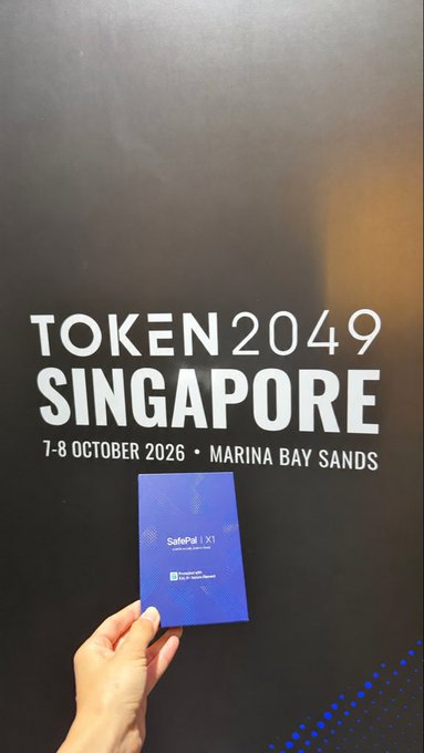A hand holding a 'SafePal X1' hardware wallet card in front of a 'TOKEN 2049 SINGAPORE' event backdrop for '7-8 OCTOBER 2026 • MARINA BAY SANDS'.