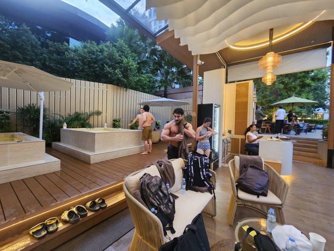 A shirtless man and others in swimwear gather in a luxurious outdoor wellness lounge featuring wooden decking, wicker seating, a hot tub, and warm lantern lighting, with backpacks and water bottles scattered about, suggesting a post-activity social mixer among the founders and builders described in the post.