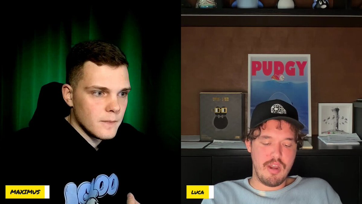 The image shows two men sitting next to each other, both wearing hats. One man has a black hat with "Fuggy" written on it, while the other man wears a blue hat. The background features a green curtain that covers part of a wall and several books arranged on shelves or a bookcase behind them.