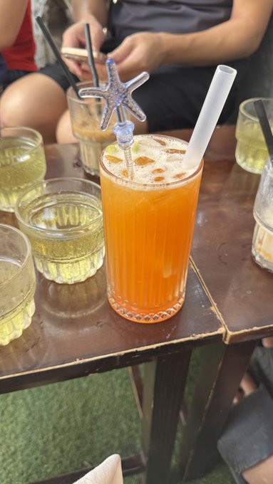 An orange iced drink with a starfish garnish and a white straw sitting on a wooden table, surrounded by other drinks.