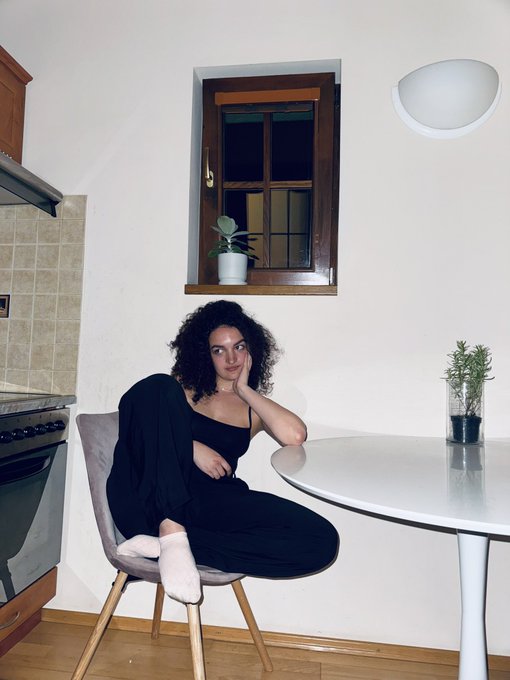 A young woman with curly hair sits casually at a white round kitchen table, wearing a black outfit and pink socks, looking directly at the camera.