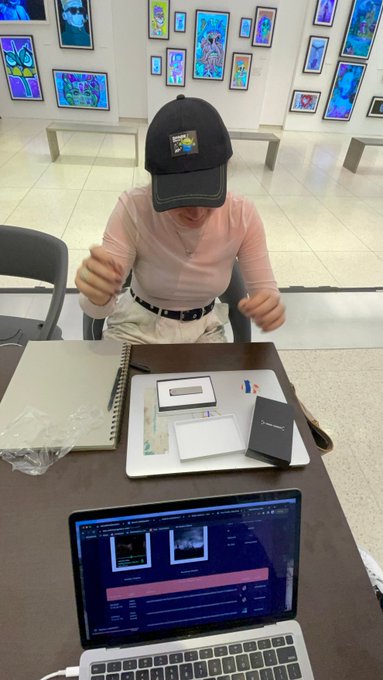 A person wearing a black cap and pink top sits at a desk working on a MacBook that displays video editing software, with packaging materials, cards, and pens scattered on the table in what appears to be an art gallery.