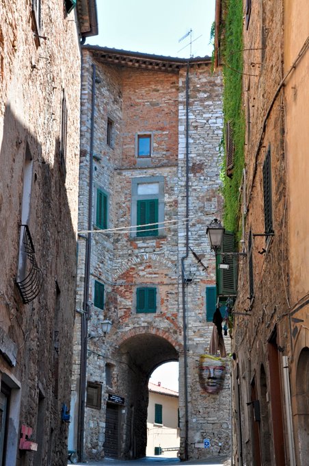 A narrow stone alleyway in a European town featuring an arched passageway, brick buildings with green shutters, ivy growing on the walls, and a decorative face sculpture mounted near the archway.