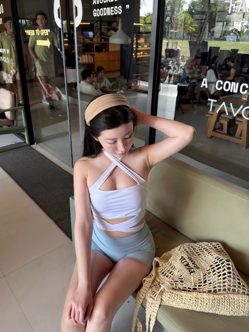 A young woman wearing a beige headband, white halter top, and light blue shorts sits on a bench outside a cafe, with a net bag beside her.