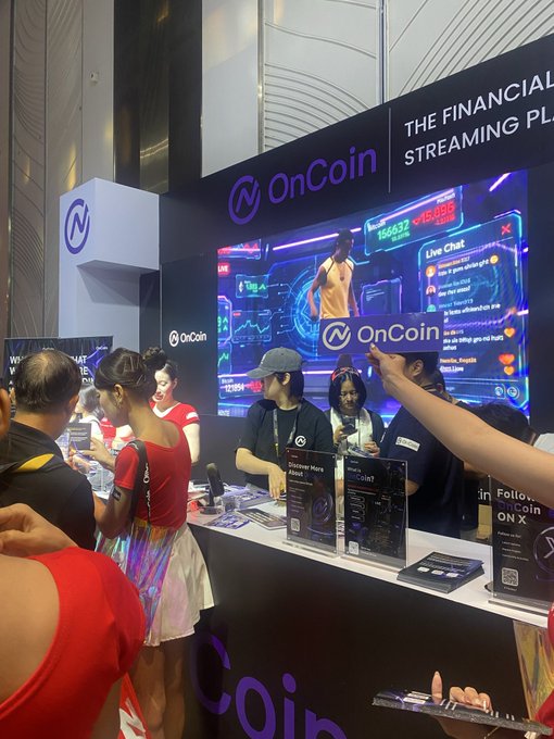 A busy booth at an exhibition for 'OnCoin' featuring 'THE FINANCIAL STREAMING PL...' signage, large screens, and people engaging with staff at the counter.