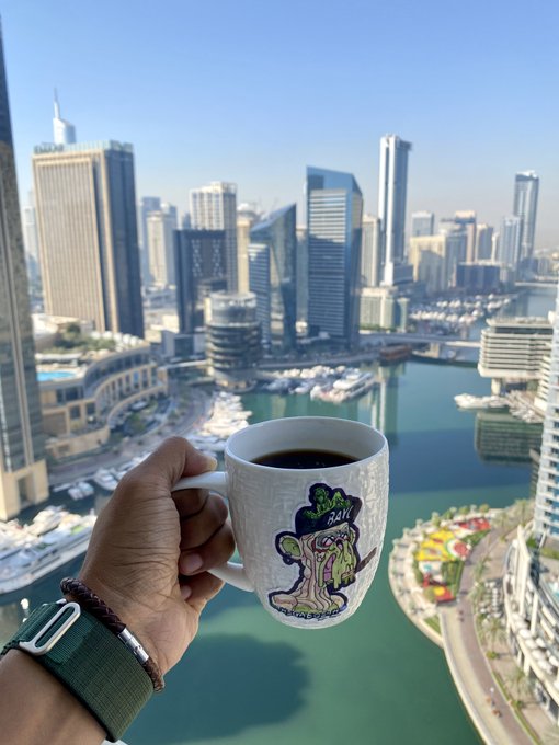 iced coffee with a green frog design, held by a hand on a balcony overlooking the Dubai skyline and the water below.