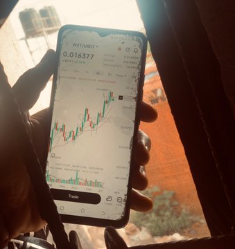 A hand holding a smartphone displaying the WXT/USDT trading pair at 0.016377 with a candlestick price chart.