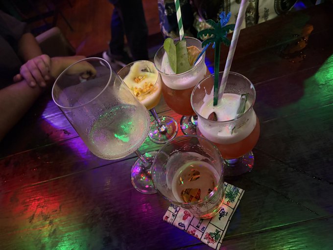 Four colorful cocktails in various glasses with straws and garnishes sit on a wooden table illuminated by colorful lights, with a person's arm visible in the background.