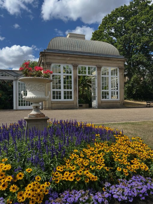 A classical stone building with a domed glass roof stands behind a flowerbed of yellow black-eyed Susans, purple salvias, and a white decorative urn filled with pink flowers.