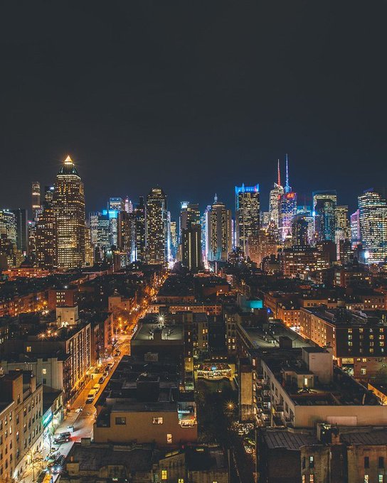 An aerial nighttime view of a sprawling city skyline featuring numerous illuminated skyscrapers and a network of glowing streets stretching into the distance.