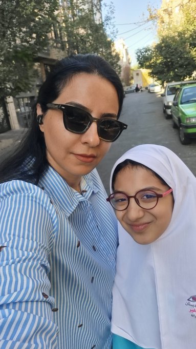 A selfie of a woman in sunglasses and a striped shirt standing next to a smiling young girl wearing a white hijab and glasses, taken outdoors on a sunny street.