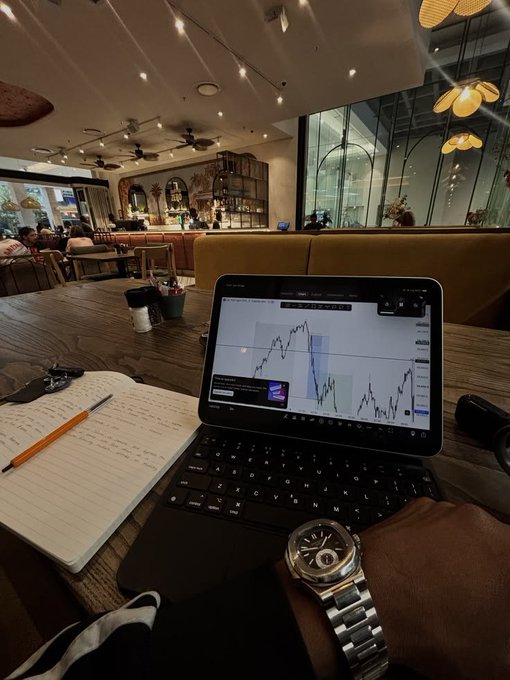A first-person view of someone using a tablet displaying a financial chart in a cafe, with a notebook, pen, and a luxury watch on their wrist.