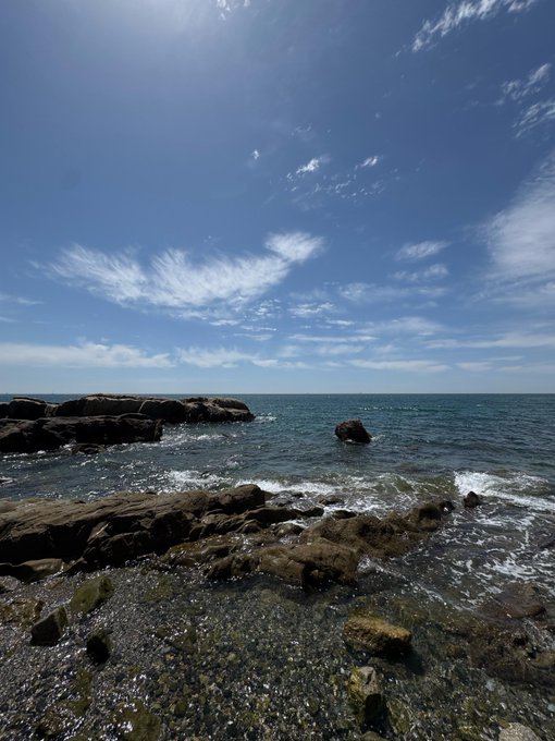 A scenic view of a rocky coastline under a bright blue sky with wispy clouds, featuring waves crashing against the shore.
