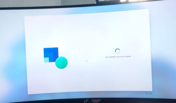 A photograph of a curved monitor displaying a Windows update screen with the message 'Just a moment, checking for updates' and a loading circle.