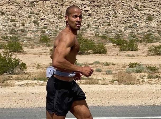 A shirtless, muscular man in black running shorts jogs along a desert road with rocky, arid terrain in the background.