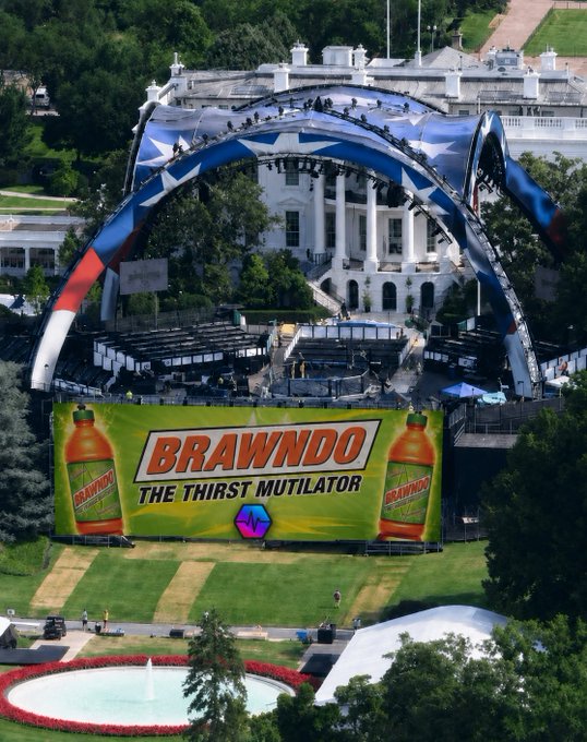 The image features a large billboard with an advertisement for Brandywine, a type of alcohol. The billboard is located outside a white building that appears to be a government facility or event venue. There are several people visible around the area, possibly attending an outdoor event or gathering. A fountain can also be seen in the vicinity, adding to the overall atmosphere of the scene.