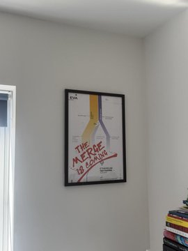 A framed movie poster for 'THE MERCENARY IS DEAD' hangs on a white wall next to a window and a stack of colorful boxes.