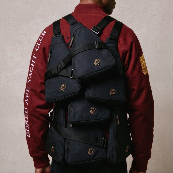 A person wearing a maroon 'BORED APE YACHT CLUB' sweatshirt models a navy tactical-style backpack with multiple flap pockets, each adorned with a small gold skull logo.