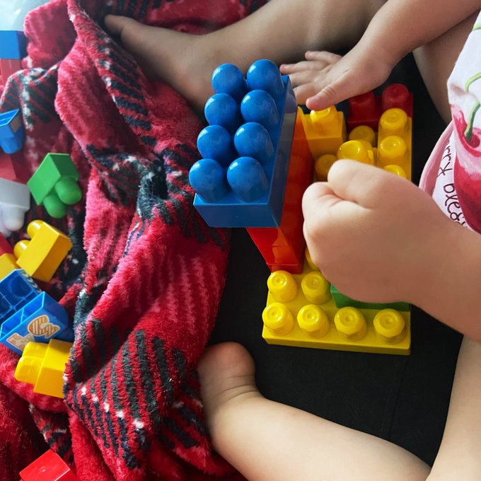 A baby plays with colorful building blocks, reaching out to touch a red block on top of a blue and yellow stack, resting on a red patterned blanket.