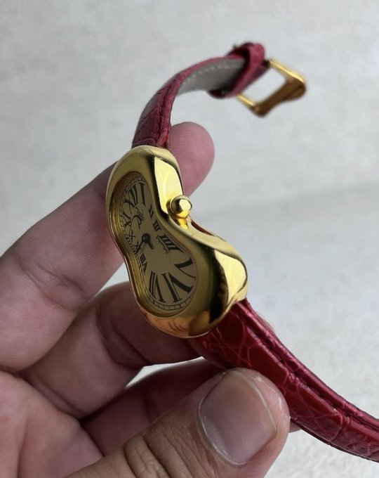 A close-up photograph of a hand holding a surreal Salvador Dalí-inspired "melting" wristwatch with a gold-toned face, distorted black Roman numerals, and a textured red leather strap.