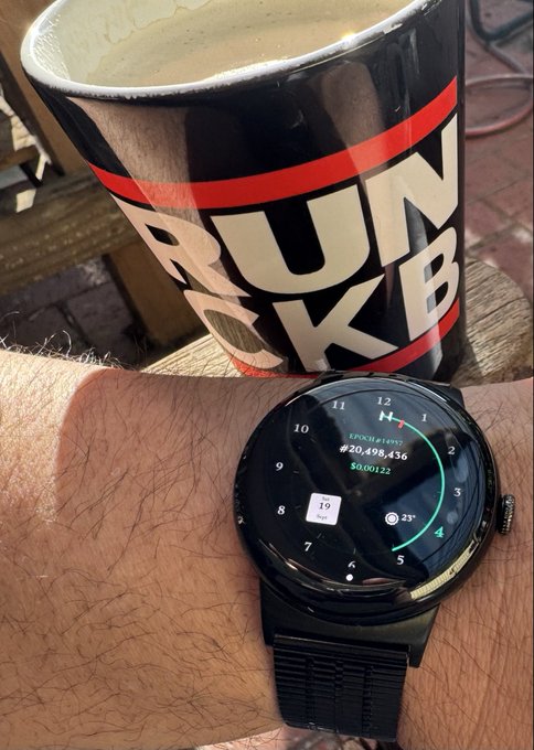 A close-up of a hairy wrist wearing a black Huawei smartwatch displaying '#20,498,436' and $0.00122, positioned next to a Dunkin' coffee mug.