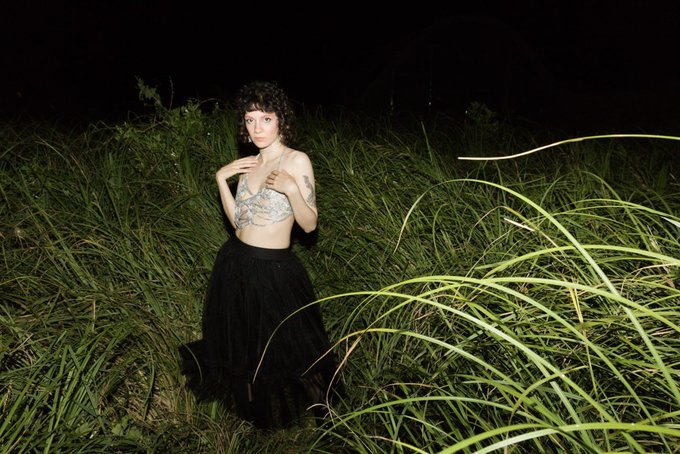 A woman with short dark curly hair, wearing a lace top and a long black skirt, stands in a field of tall grass at night.
