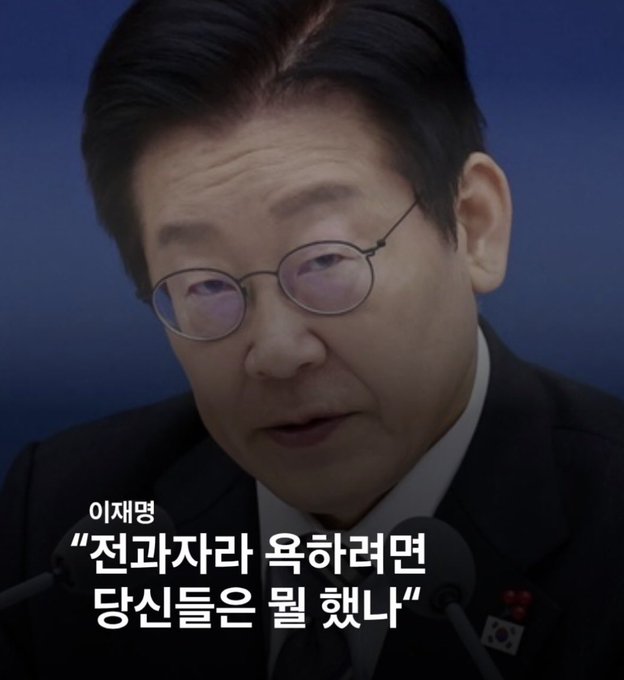 The image shows a man with glasses speaking into microphones, with Korean text overlaid reading "이재명 '전과자라 욕하려면 당신들은 뭘 했나'" (Lee Jae-myung: "If you're going to criticize me for having a criminal record, what have you done?"). The caption "Unlike you, I lived fucking hard without any priors. But what the hell are you staring at?" appears to be a crude English reinterpretation or parody of Lee Jae-myung's defiant message challenging his critics, though it introduces vulgar language not present in the original Korean quote.