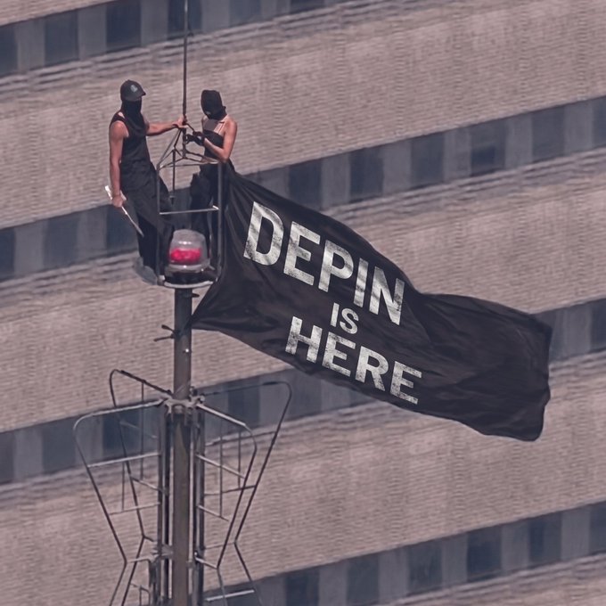 Two masked individuals sit atop a tall metal pole or tower, unfurling a large black flag that reads 'DEPIN IS HERE' against a backdrop of a stadium structure.