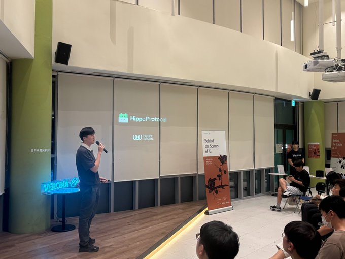 A person speaks into a microphone at a presentation in a modern venue, with a 'Median' neon sign and logos for 'Hippoprotocol' and 'GO! DRIZZ DRIZZ' visible, alongside a banner reading 'Beyond the Scenes of AI'.