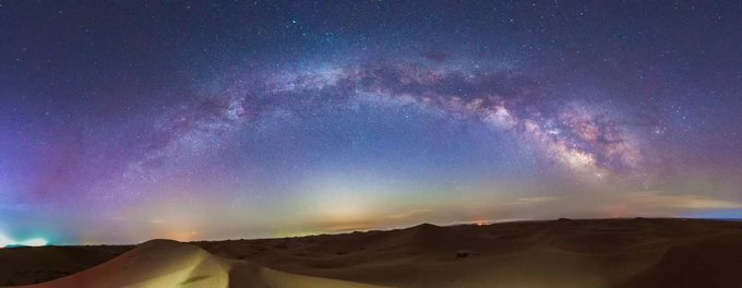 The Milky Way arches brilliantly over a vast expanse of dark desert dunes, while faint aurora-like lights glow on the horizon—almost as if the photographer has revealed a hidden, otherworldly "cutscene" in nature's grandest display.