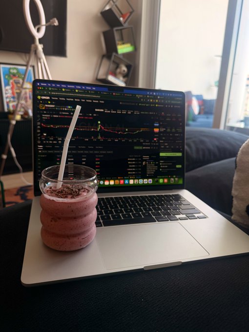 A laptop displaying a stock trading chart sits on a couch next to a pink smoothie topped with chocolate shavings in a ribbed glass.
