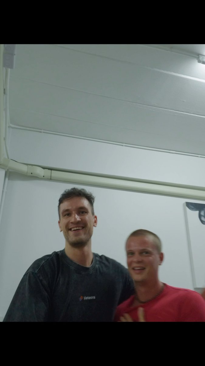 Two men stand close together indoors; one wears a dark shirt with 'Meteora' text and smiles at the camera, while the other wears a red shirt and smiles beside him.