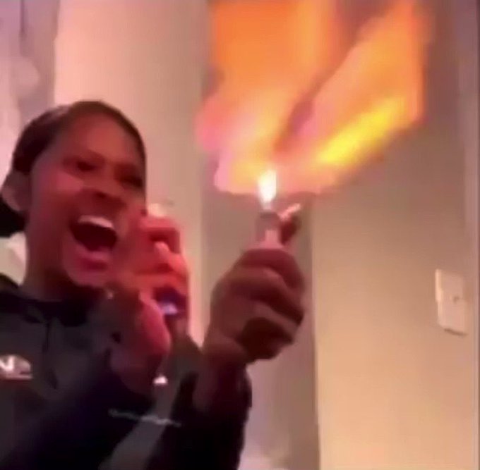 The image shows a woman holding a lit lighter, with her mouth open as if she's making a funny or surprising face while talking on her cell phone. The scene appears to be set against a white wall, which contrasts with the bright flame from the lighter. The background is blurred and out of focus, drawing attention to the main subjects in the image - the woman holding the lit lighter and herself engaged in conversation on the phone.